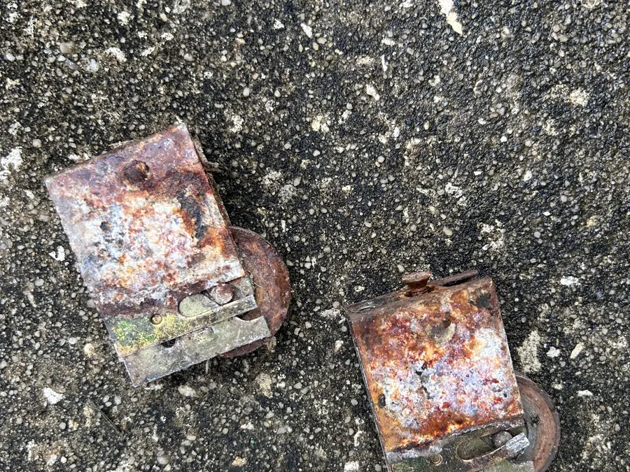 Two sliding door roller assemblies almost entirely consumed by rust, lying on asphalt.